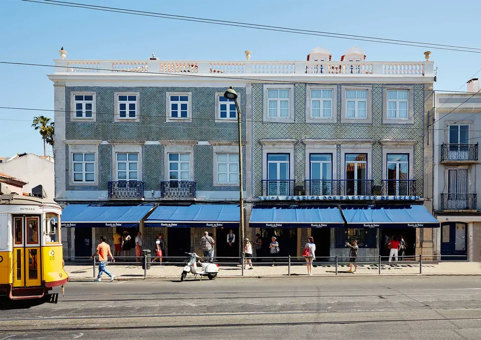 La façade carrelée et les auvents bleus de la maison Pastéis de Belém, un tramway jaune passant devant